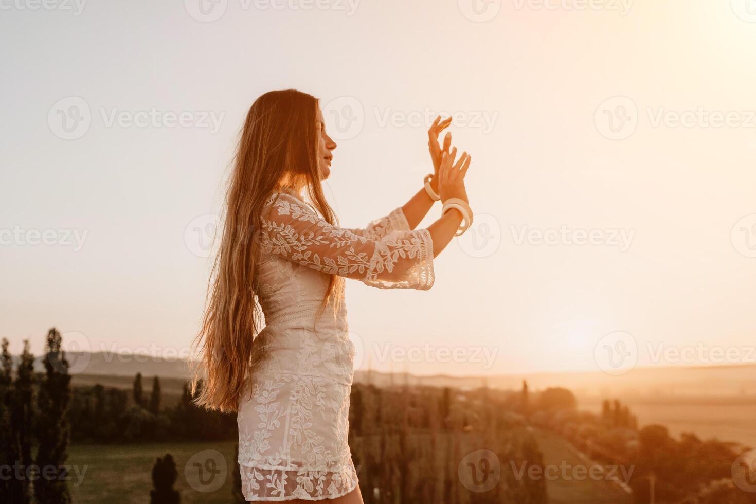 Happy woman in white boho dress on sunset in mountains. Romantic woman with long hair standing with her back on the sunset in nature in summer with open hands. Silhouette. Nature. Sunset. photo