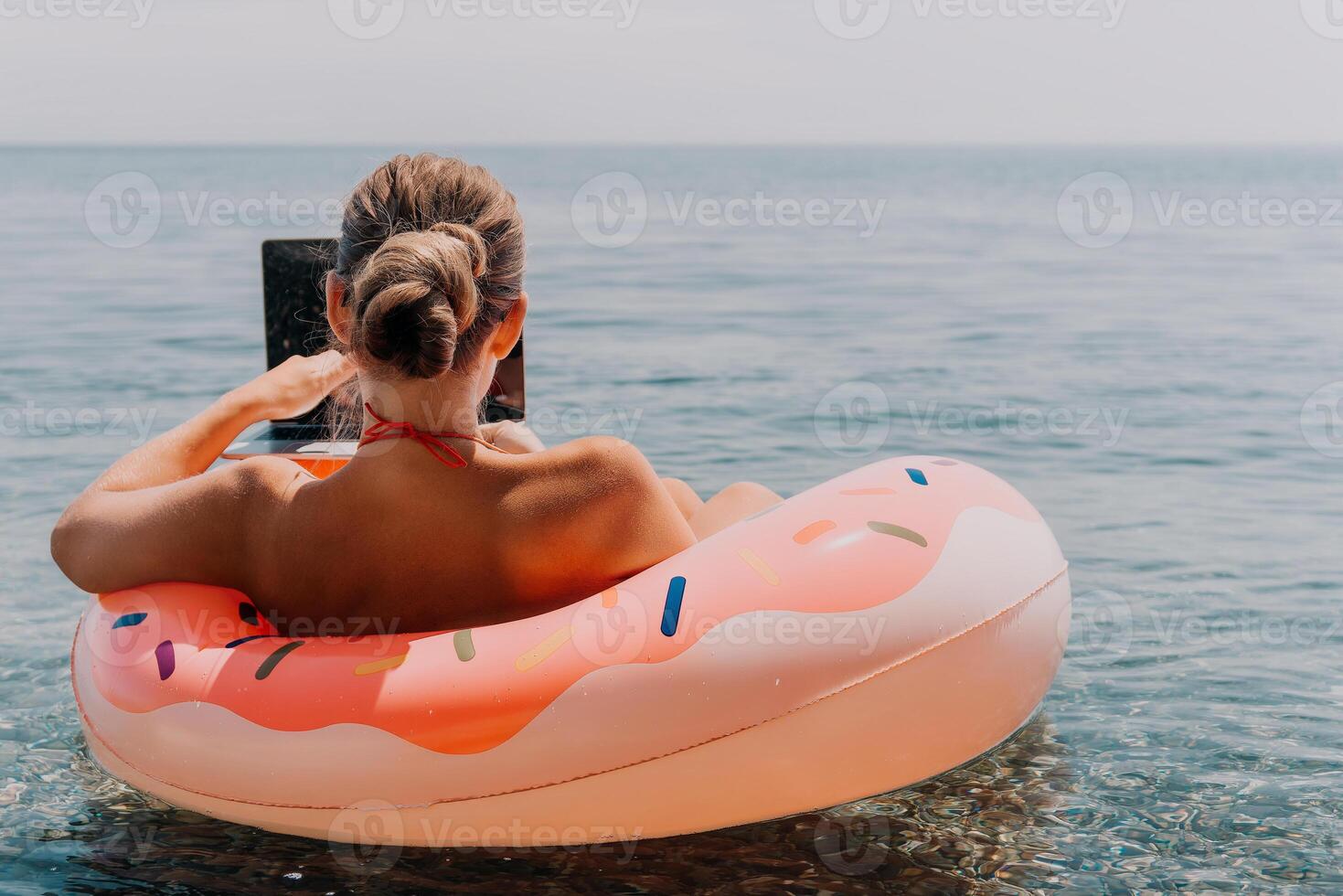 Woman freelancer works on laptop swimming in sea on pink inflatable ring. Happy tourist floating on inflatable donut and working on laptop computer in calm ocean. Freelance, remote working anywhere photo