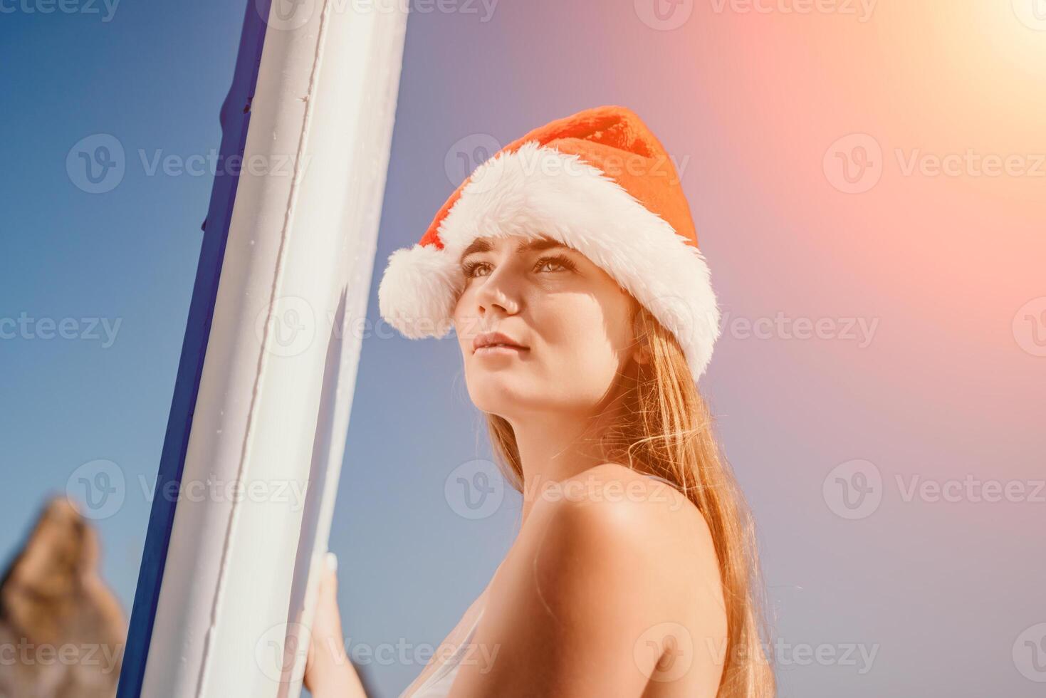 Woman sea sup. Close up portrait of happy young caucasian woman with long hair in Santa hat looking at camera and smiling. Cute woman portrait in a white bikini posing on sup board in the sea photo