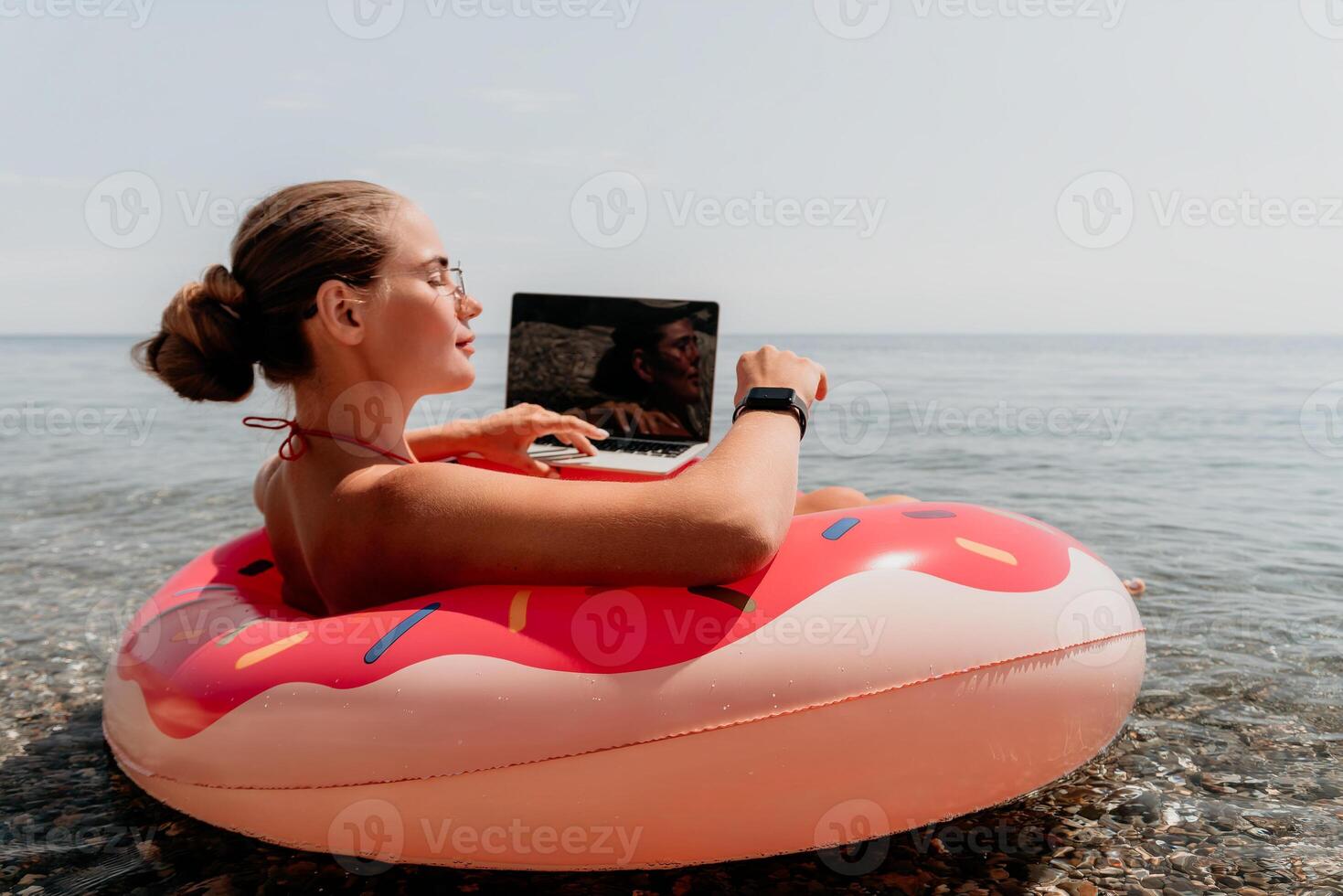Woman freelancer works on laptop swimming in sea on pink inflatable ring. Happy tourist floating on inflatable donut and working on laptop computer in calm ocean. Freelance, remote working anywhere photo
