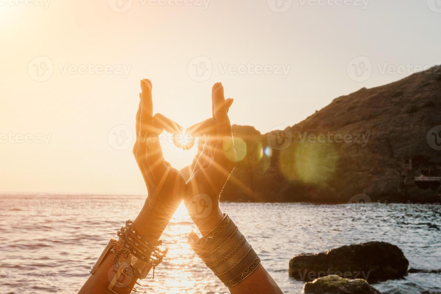 Woman sea yoga. Happy woman in a white swimsuit and boho-style bracelets making a heart shape with her hands while practicing yoga on a mat by sea at sunset. Healthy lifestyle, harmony and meditation photo
