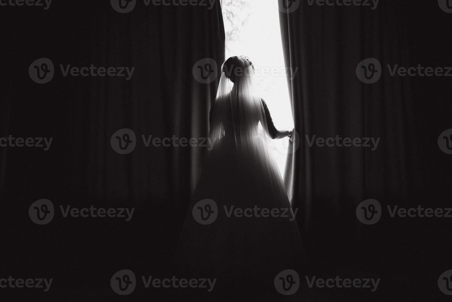 Portrait of beautiful bride in white silk dressing gown with curly hairstyle and veil standing near window in bedroom, copy space. Bride's morning preparation. Back view photo