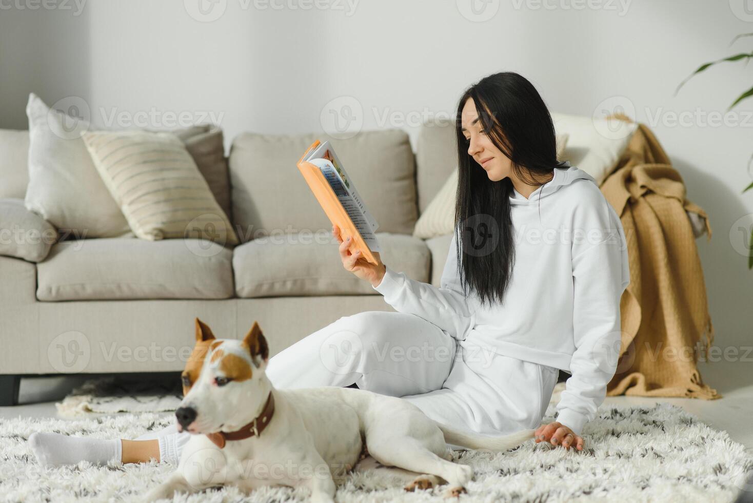 Young smiling woman resting on sofa and reading a book with dog sitting near by her at home. photo