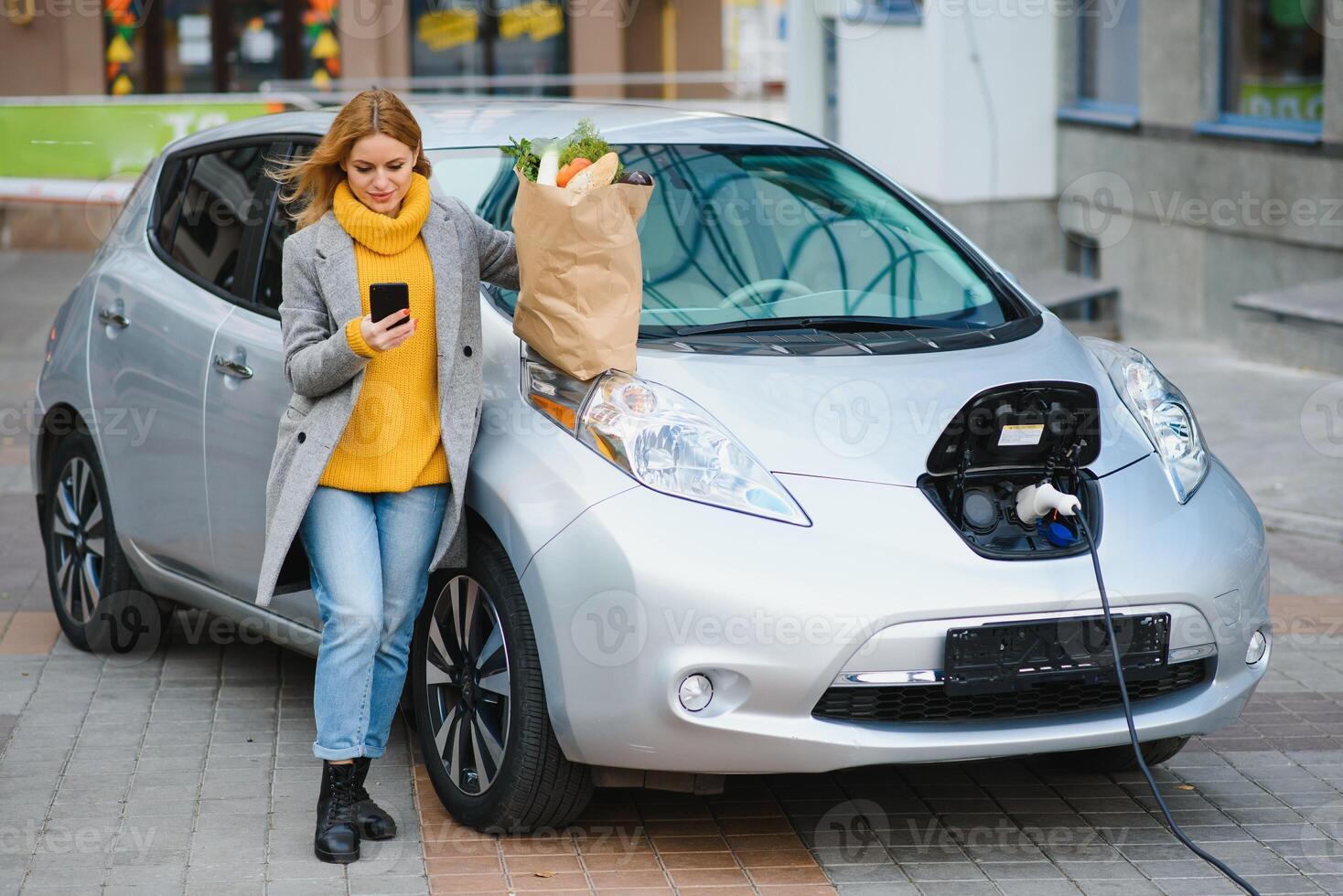 Using smartphone while waiting. Woman on the electric cars charge station at daytime. Brand new vehicle photo
