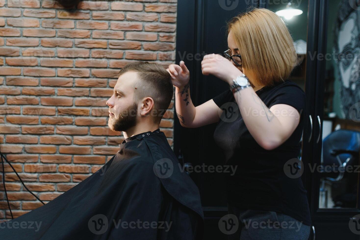 Serving client in Barbershop. Professional barber girl, female hairdresser making modern haircut for a man sitting in barber shop chair. Focus on a girl. Hairdressing, shaving, trimming, grooming photo