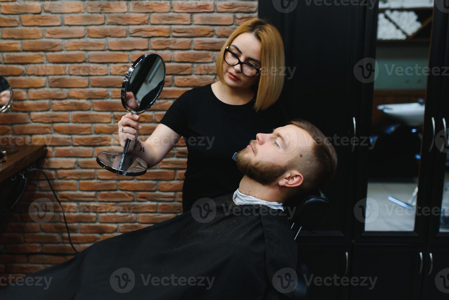 Young adult beautiful caucasian woman hairdresser cuts beard handsome man at modern barbershop Happy gay sitting chair men beauty salon. beard care. metrosexual. Female barber in black gloves serving photo