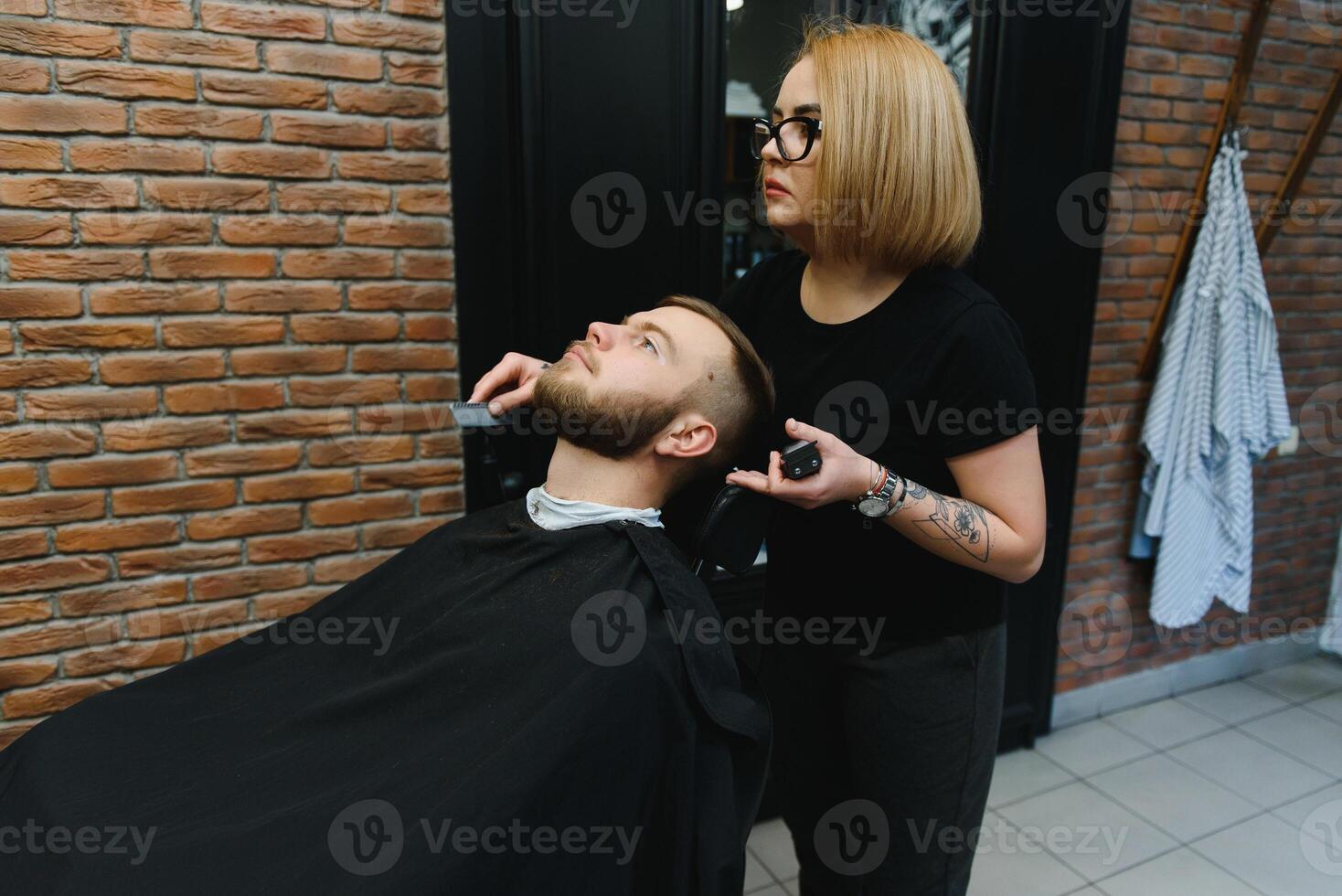 Skilled female barber cutting a beard of brunette adult guy in professional retro barbershop. photo