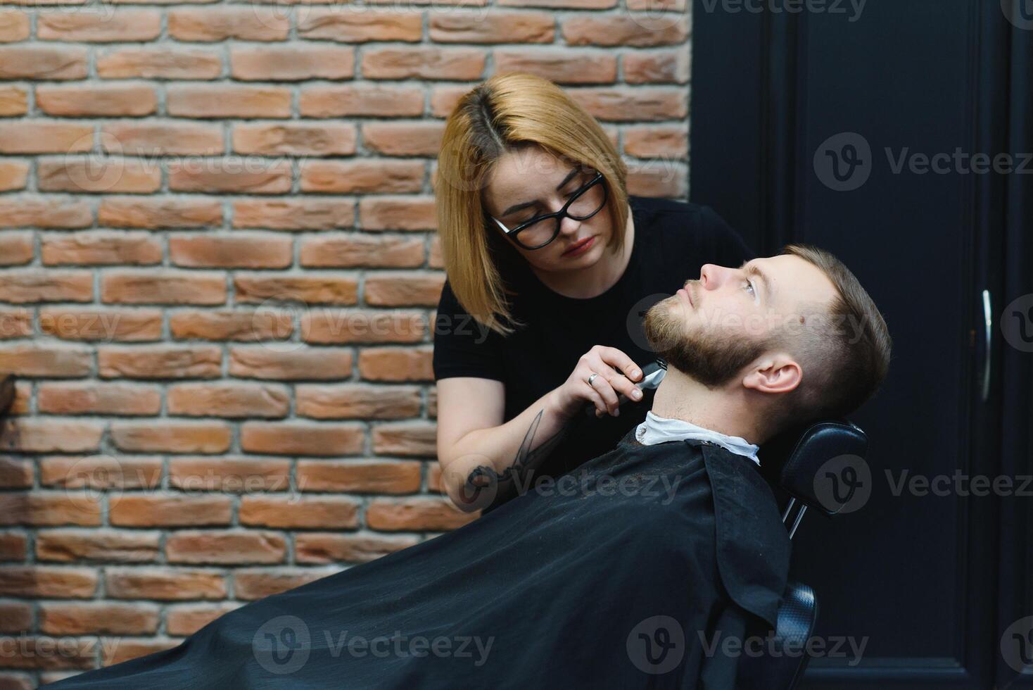 Young adult beautiful caucasian woman hairdresser cuts beard handsome man at modern barbershop Happy gay sitting chair men beauty salon. beard care. metrosexual. Female barber in black gloves serving photo