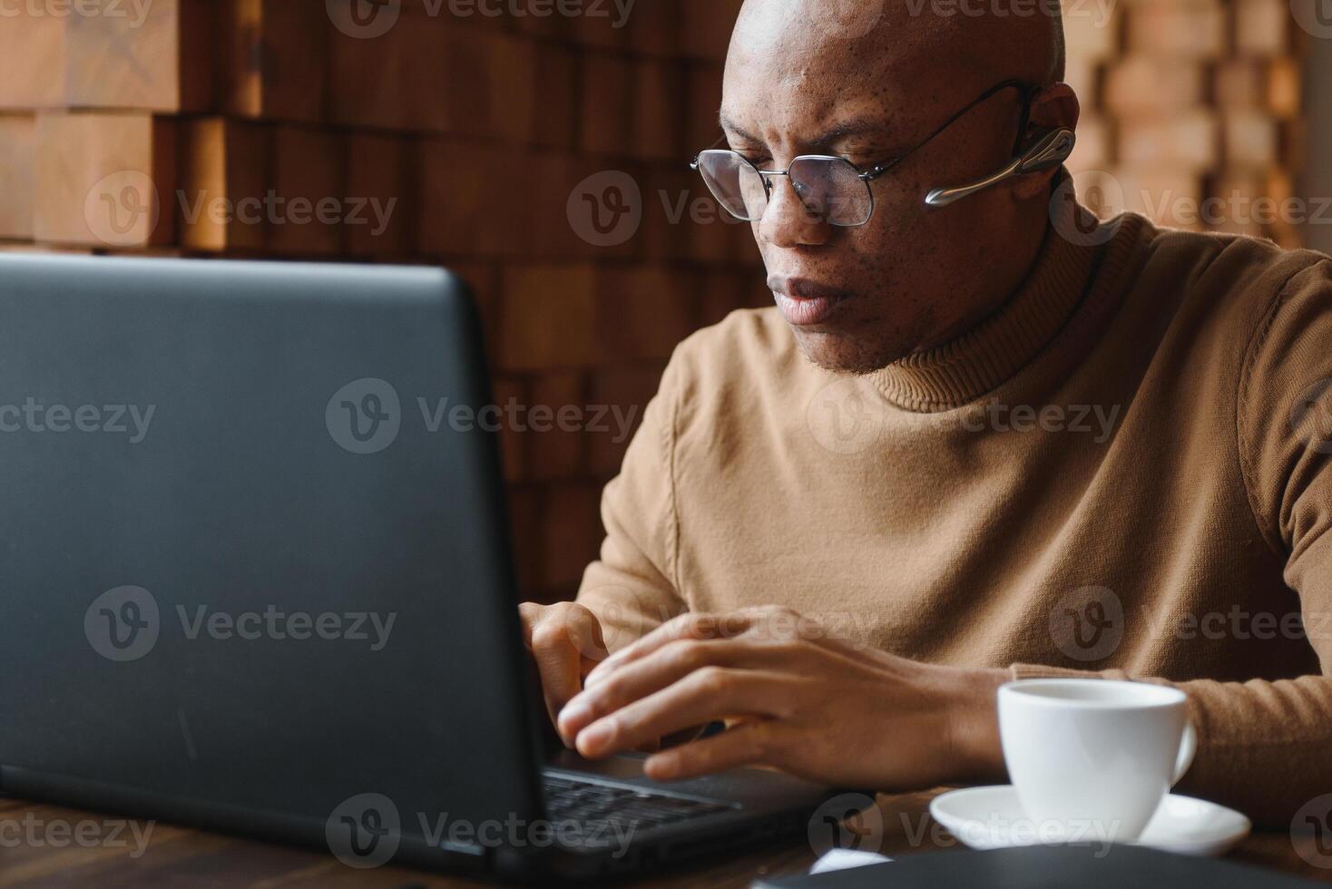 Confident prosperous African-American young top-manager wearing formal shirt having coffee and doing paperwork, working on generic laptop, using free wireless internet connection at restaurant. photo