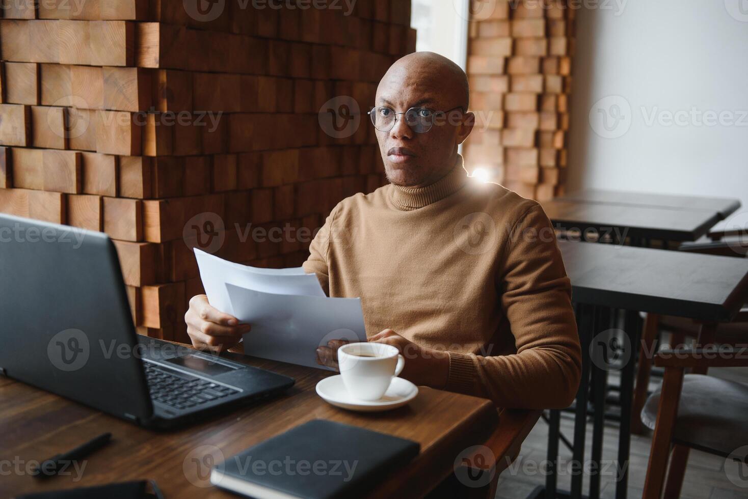 Confident prosperous African-American young top-manager wearing formal shirt having coffee and doing paperwork, working on generic laptop, using free wireless internet connection at restaurant. photo