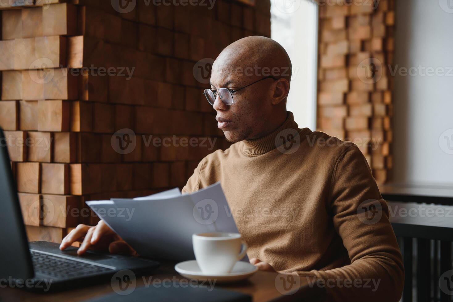 Confident prosperous African-American young top-manager wearing formal shirt having coffee and doing paperwork, working on generic laptop, using free wireless internet connection at restaurant. photo
