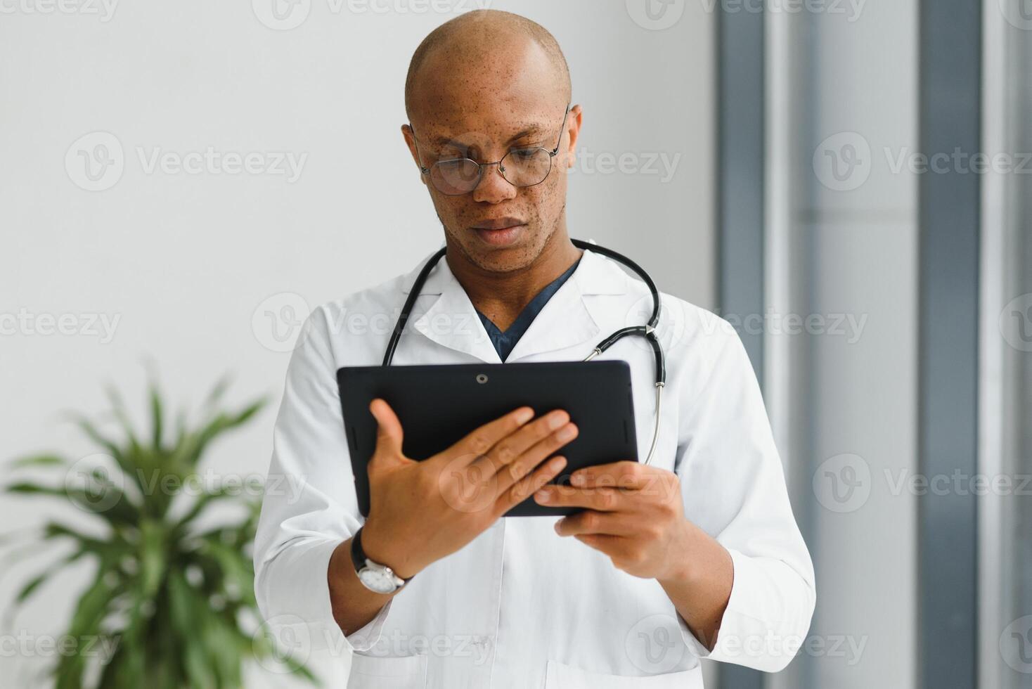 Mature african doctor using digital tablet in corridor . Portrait of confident male doctor using tablet computer in clinic with copy space. Successful smiling doctor in labcoat wearing stethoscope photo