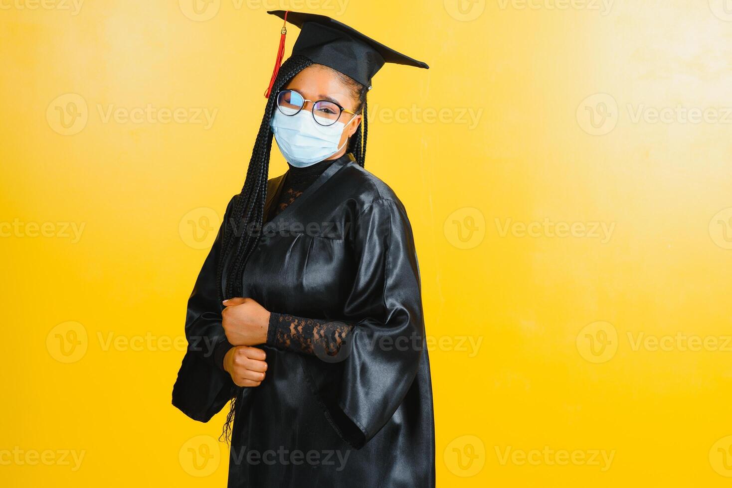 Portrait of a young african student in a graduate cap protective mask, on a yellow background, Graduation 2021 photo