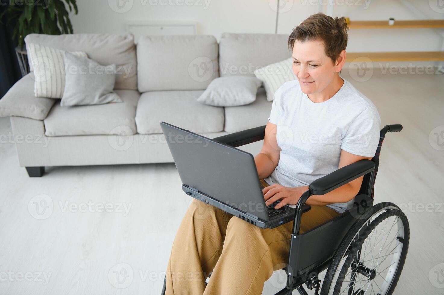 Middle age woman using laptop sitting on wheelchair at home photo