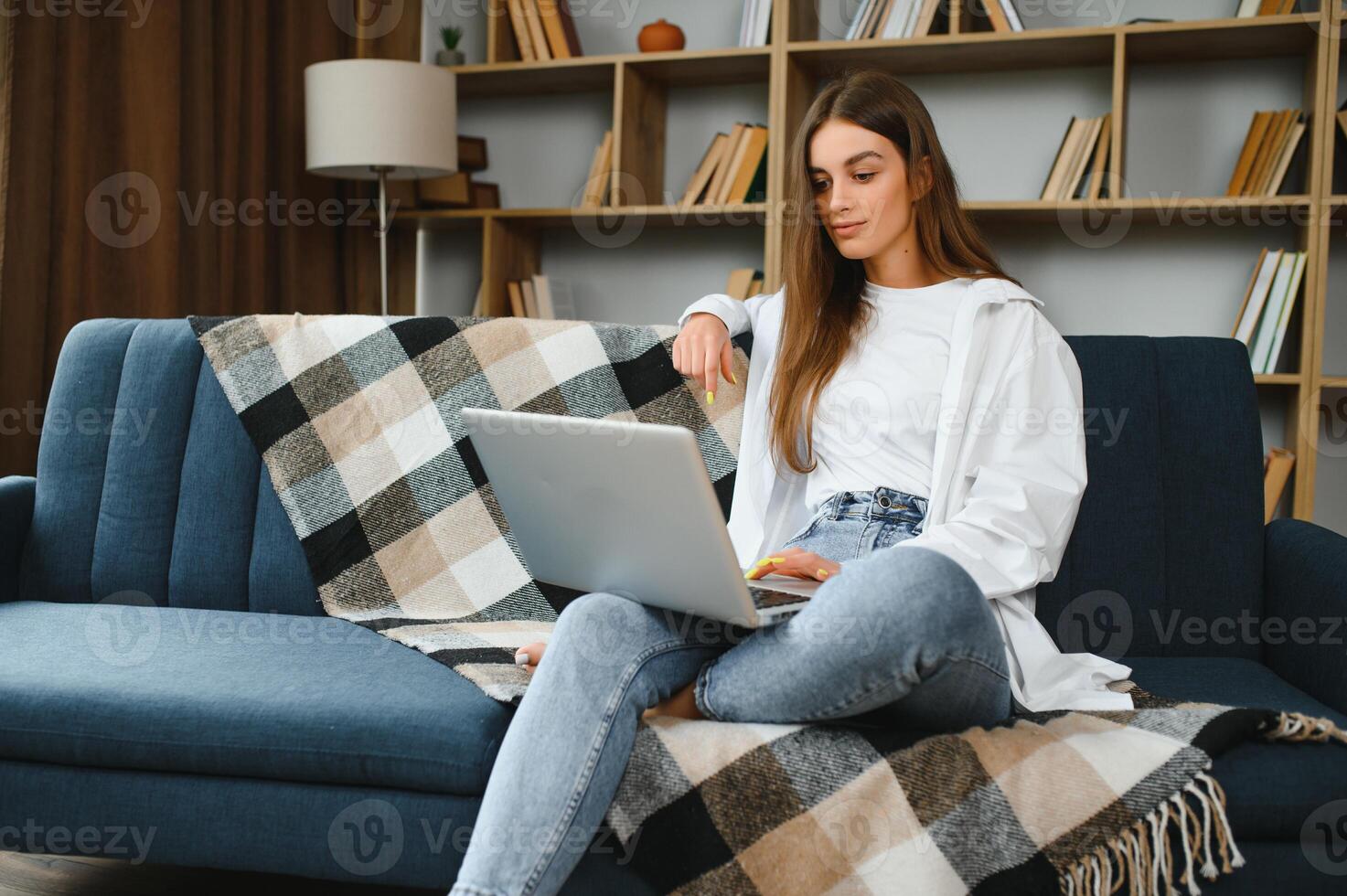 Smiling attractive young woman sitting on sofa using laptop communicating working online at home, happy teen girl typing on computer, enjoying writing blog or chatting with friends in social network photo