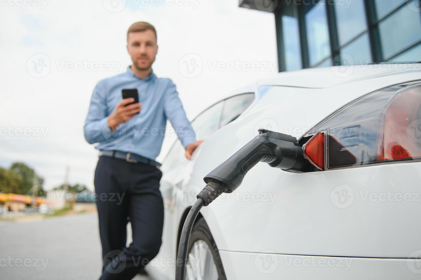 Handsome man in business suit surfing internet on modern smartphone while waiting electric car to charge. photo