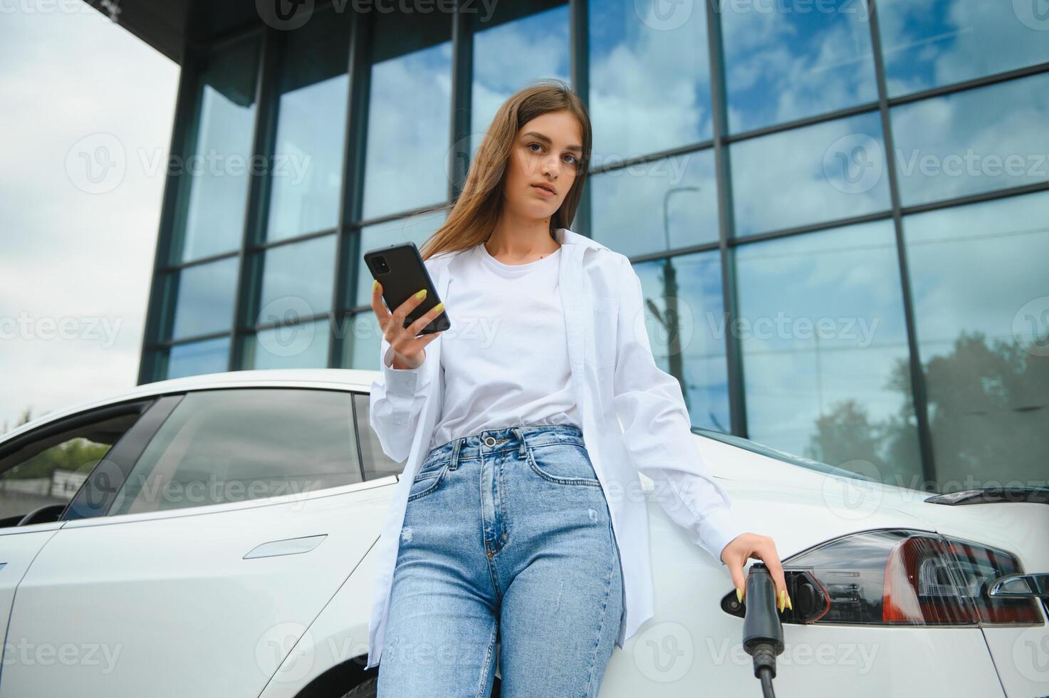 Phone in hands. Woman on the electric cars charge station at daytime. Brand new vehicle. photo