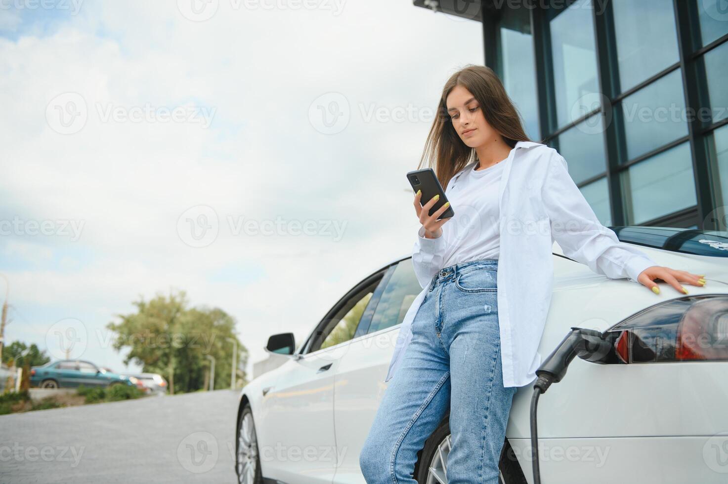 Happy young adult woman smiling wide, looking away, charging automobile battery from small public station, standing near electric car. photo