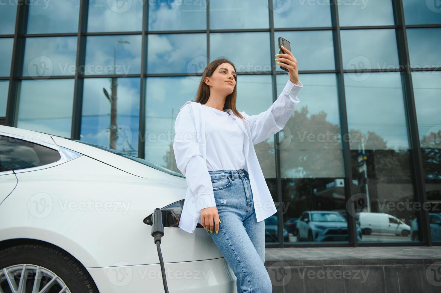 Happy young adult woman smiling wide, looking away, charging automobile battery from small public station, standing near electric car. photo
