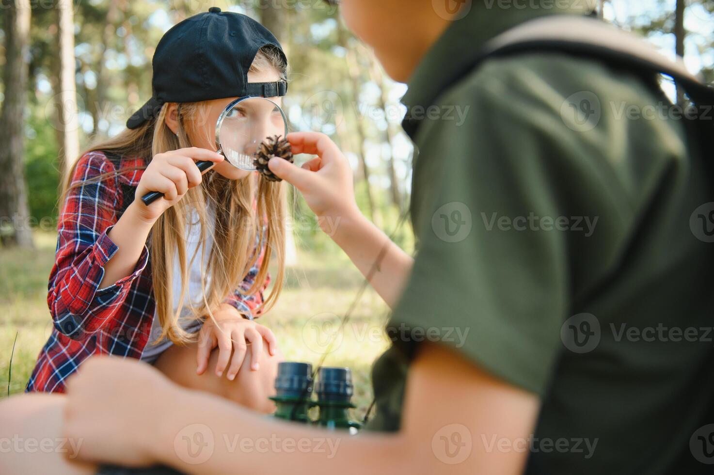Group of curious happy school kids in casual clothes with backpacks exploring nature and forest together on sunny autumn day, girl holding magnifying glass and looking at fir cone in boy hands. photo