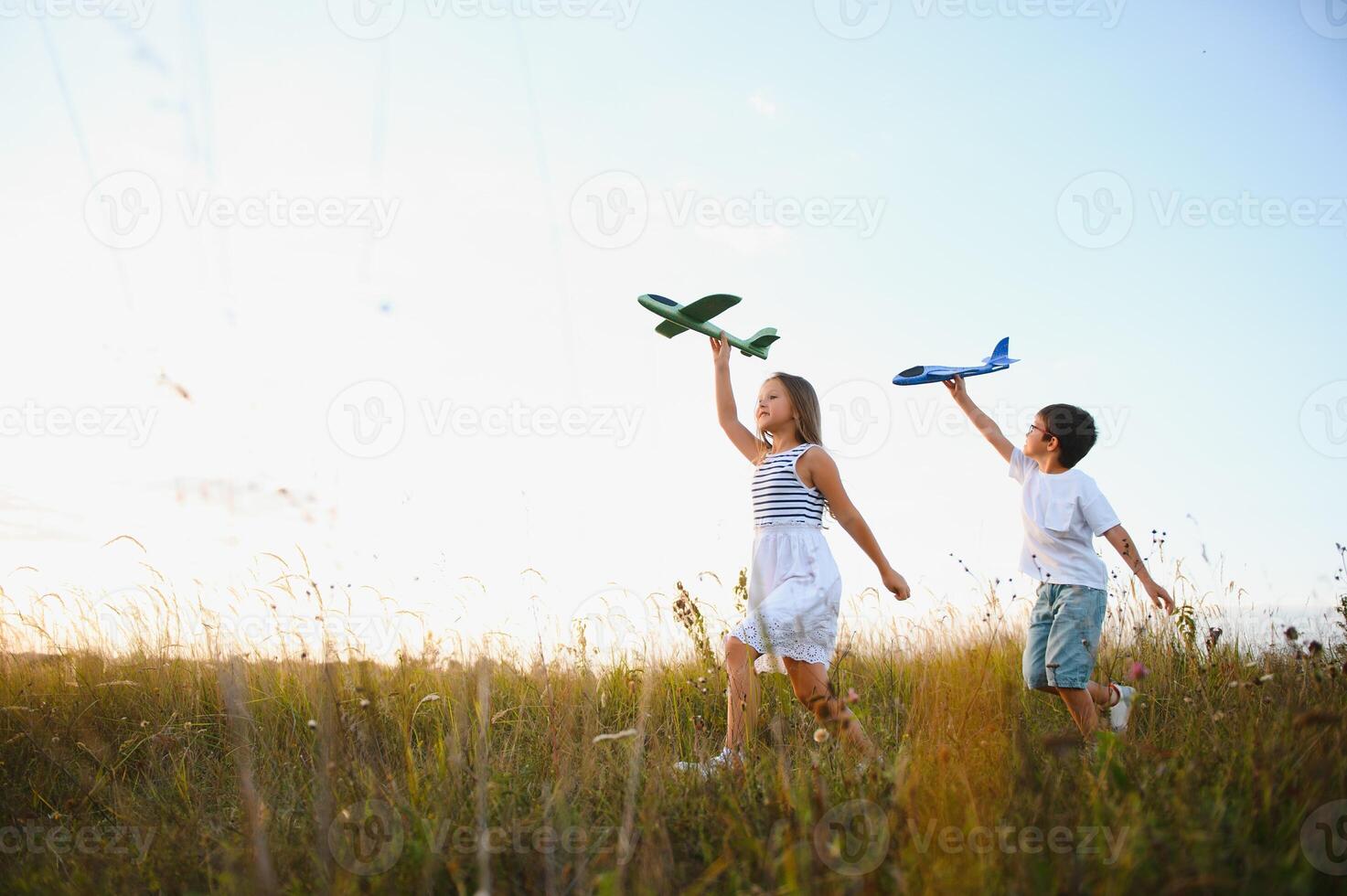 Running boy and girl holding two green and blue airplanes toy in the field during summer sunny day photo