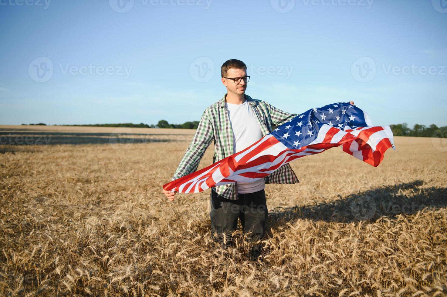 Young patriotic farmer stands among new harvest. Boy walking with the american flag on the wheat field celebrating national independence day. 4th of July concept. photo