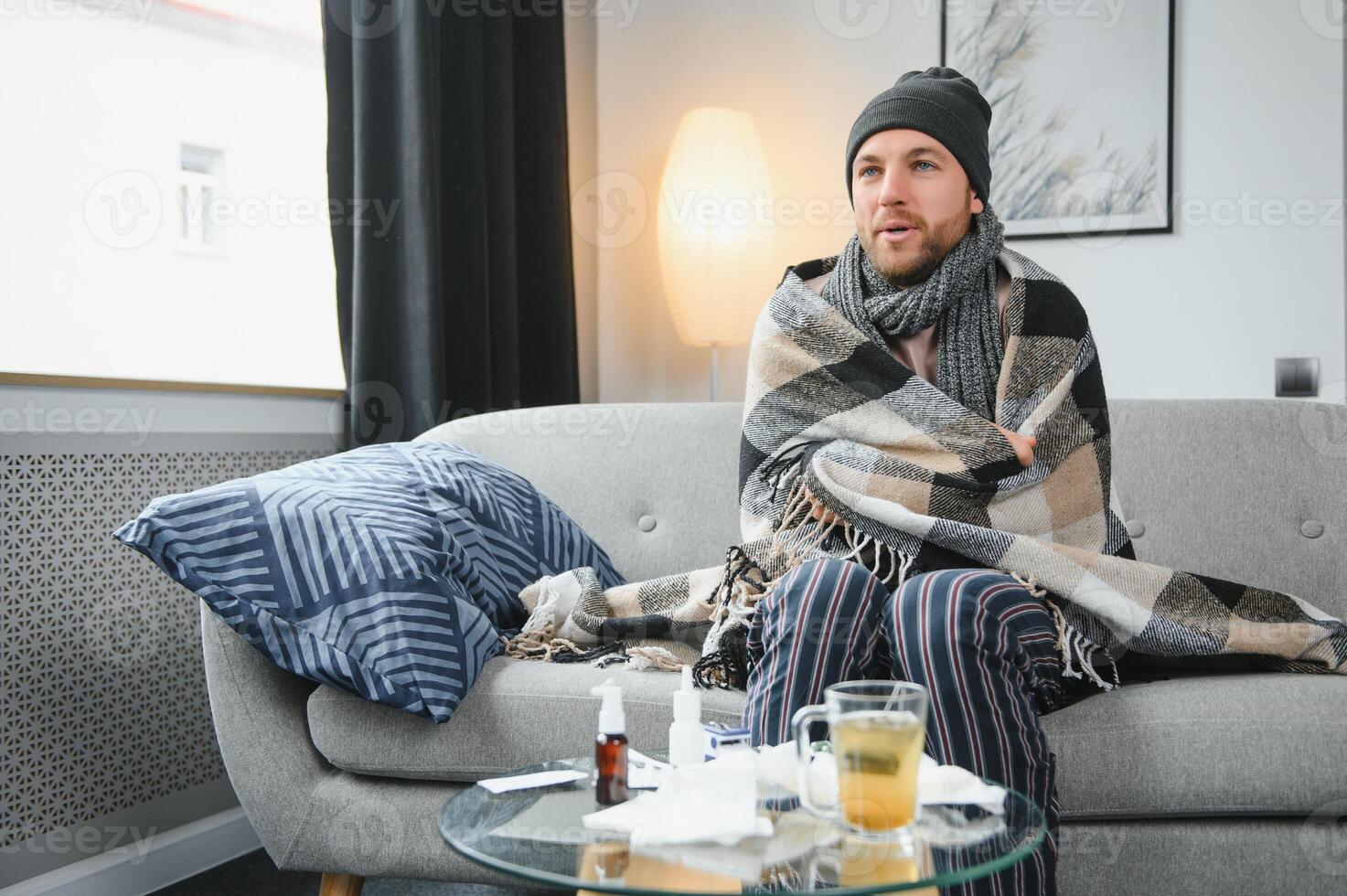 Sick bearded man who has bad cold or seasonal flu sitting on couch at home. Guy with fever wearing warm plaid shivering with worried face expression. photo