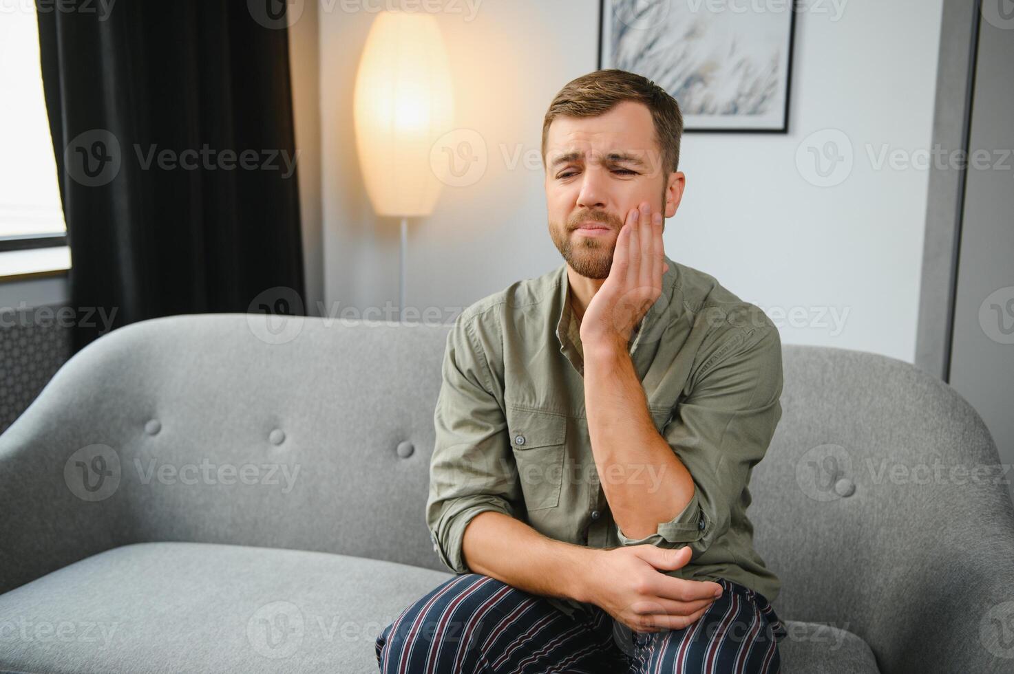 Problems with teeth. Toothache. Upset caucasian guy is sitting on the couch at home, holding his hand to his cheek, having a toothache, needs a dentist consultation, sad facial expression. photo