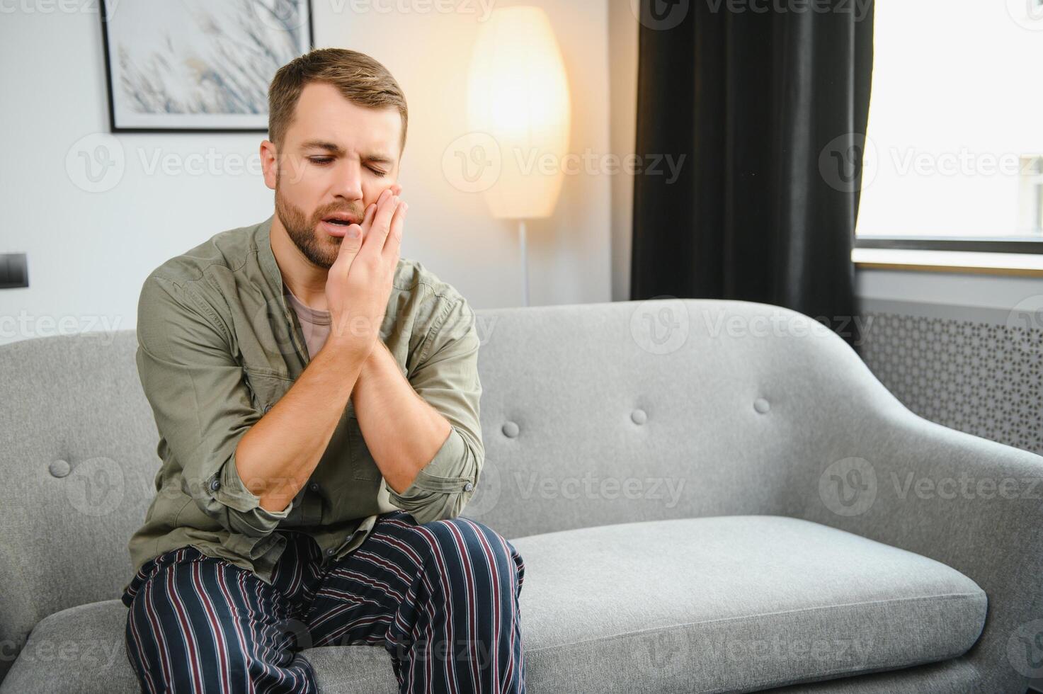 Problems with teeth. Toothache. Upset caucasian guy is sitting on the couch at home, holding his hand to his cheek, having a toothache, needs a dentist consultation, sad facial expression. photo