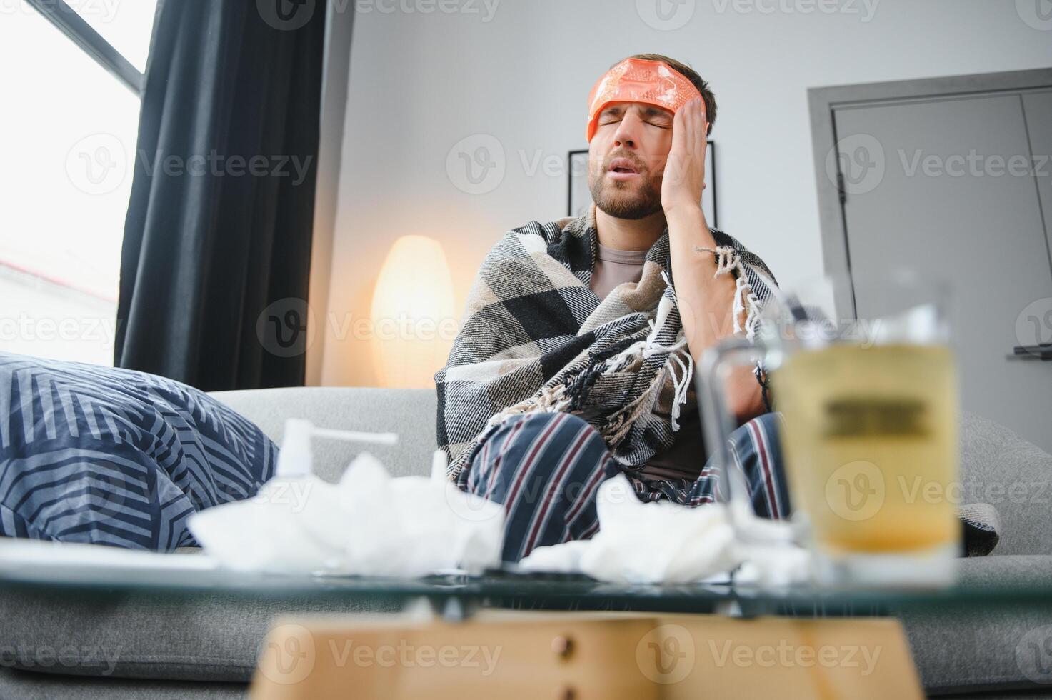 Sick bearded man who has bad cold or seasonal flu sitting on couch at home. Guy with fever wearing warm plaid shivering with worried face expression. photo