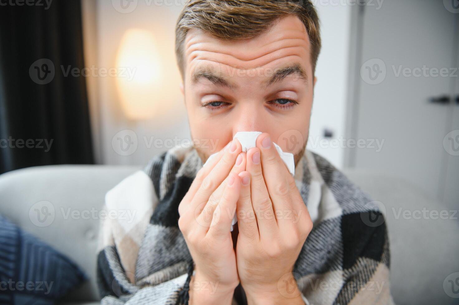 Young man suffering from a common cold and flu or allergy sit at home wrapped in blanket and wipe his nose with tissues while he have a strong headache pain, healthcare concept. photo