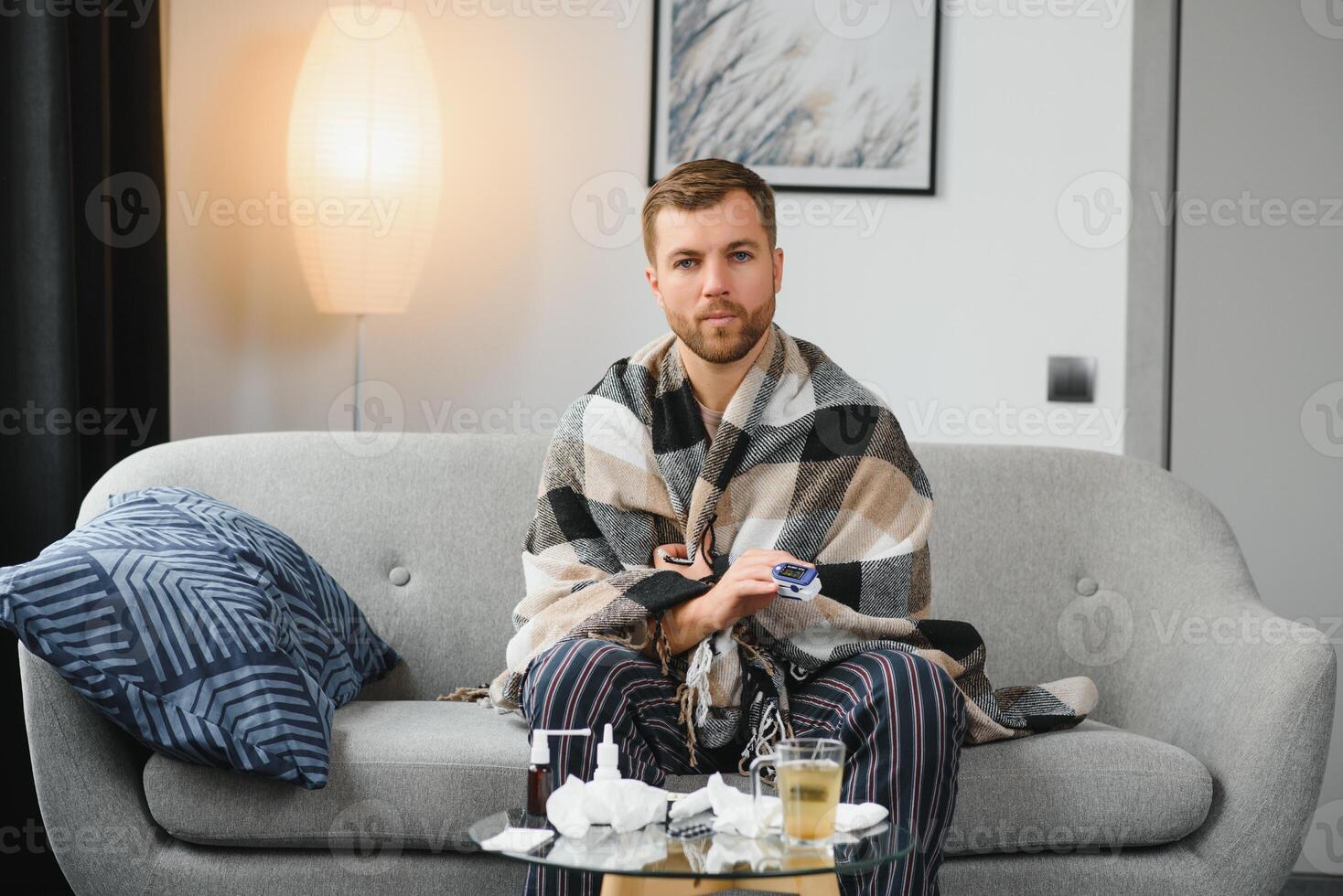 Sick adult man who has bad cold or seasonal flu virus is fighting illness at home. Unhappy middle aged man is sitting on sofa wrapped in warm scarf and plaid, holding paper tissue and sneezing. photo