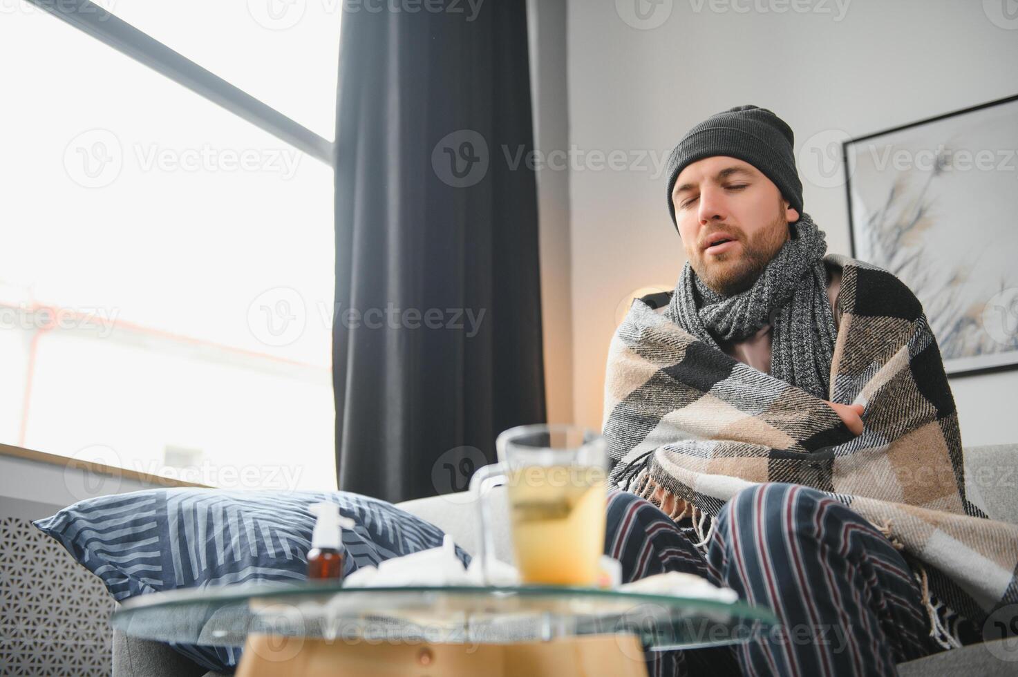 It's cold at home in wintertime. Man freezing in his house in winter because of broken thermostat. young guy wrapped in woolen plaid shivering while sitting on sofa in living room interior photo