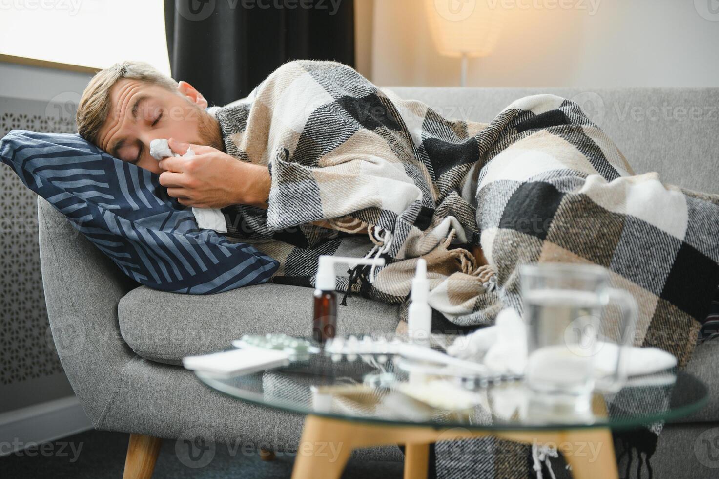 Young man suffering from a common cold and flu or allergy sit at home wrapped in blanket and wipe his nose with tissues while he have a strong headache pain, healthcare concept. photo