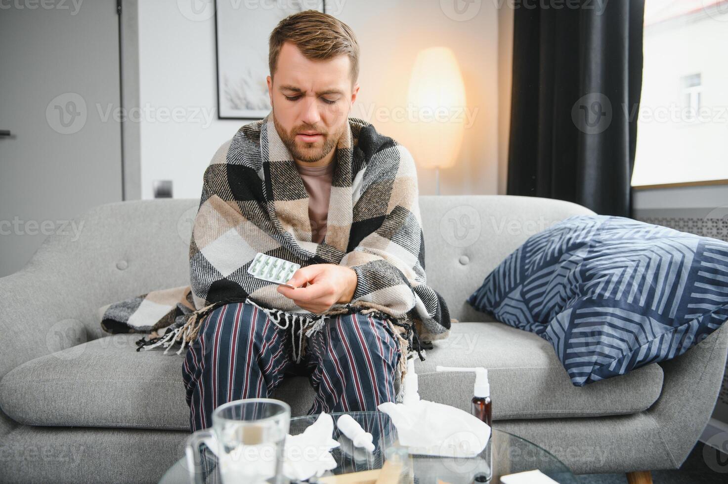 Sick bearded man who has bad cold or seasonal flu sitting on couch at home. Guy with fever wearing warm plaid shivering with worried face expression. photo