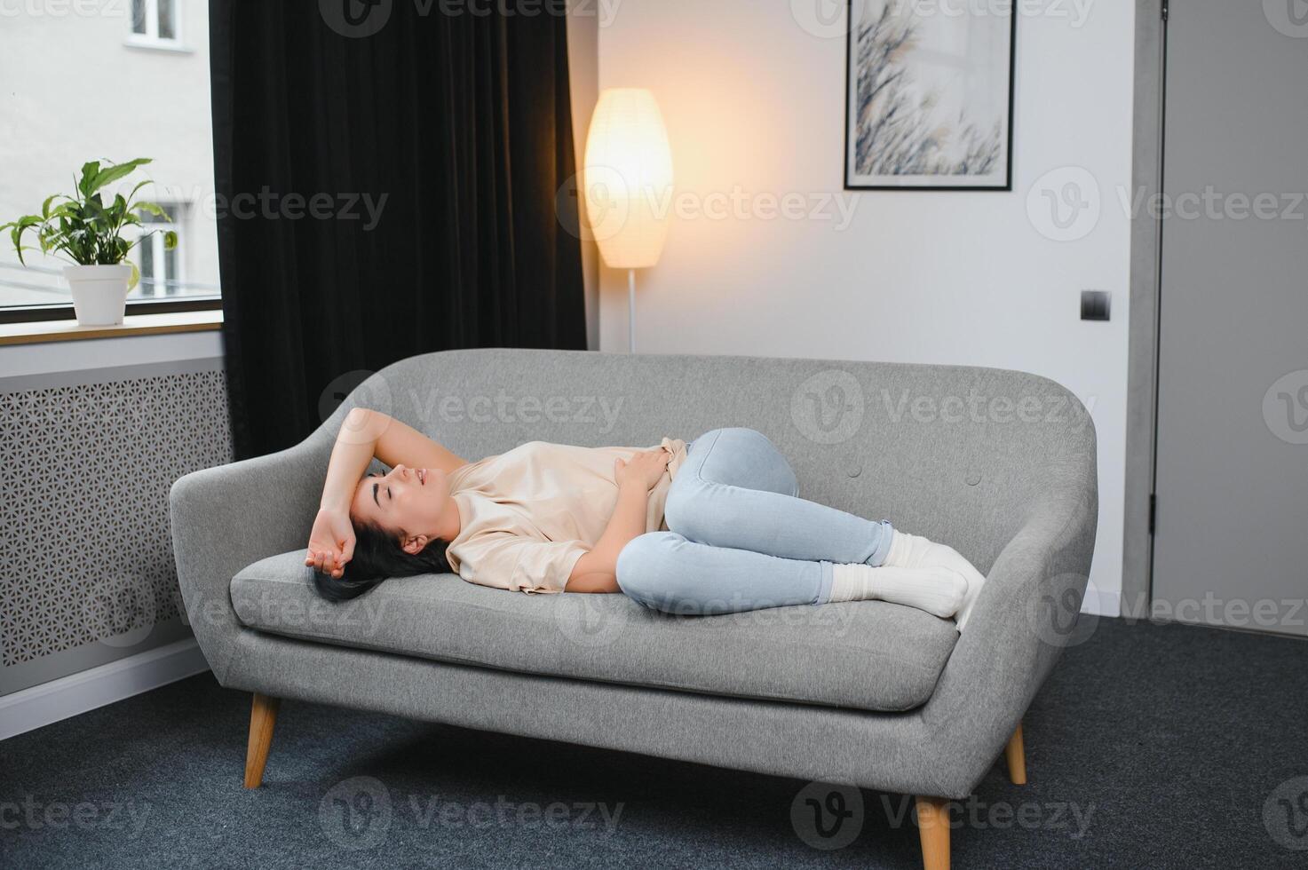 young beautiful hispanic woman in painful expression holding her belly suffering menstrual period pain lying sad on home bed having tummy cramp in female health concept. photo