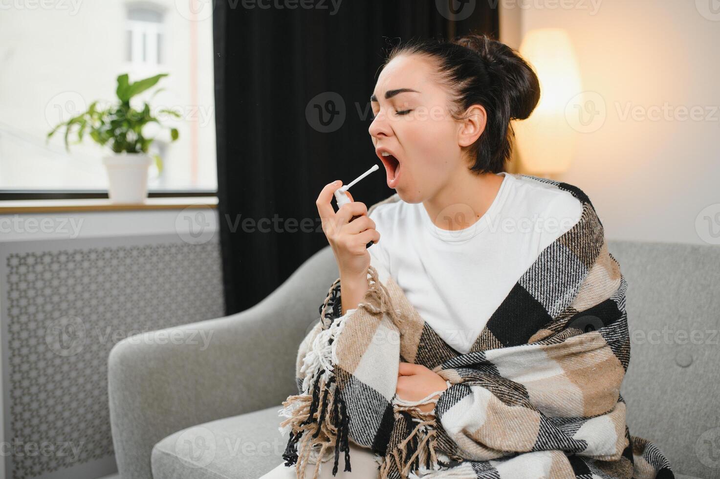 health, cold and people concept - sad sick young woman in blanket using oral spray medicine at home photo