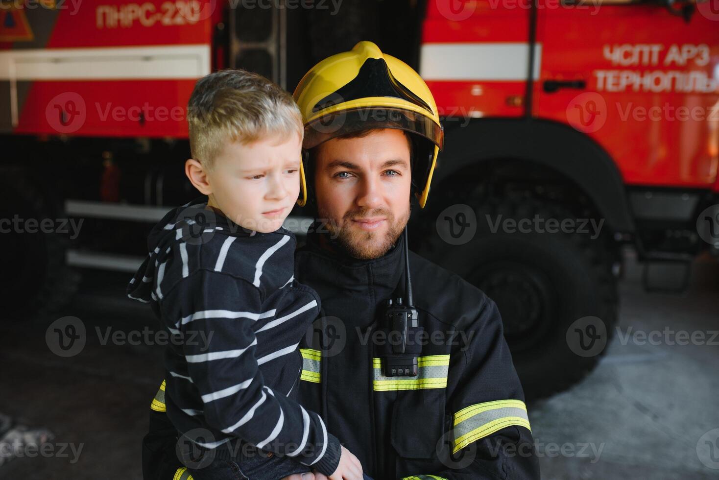 Dirty firefighter in uniform holding little saved boy standing on black background. photo