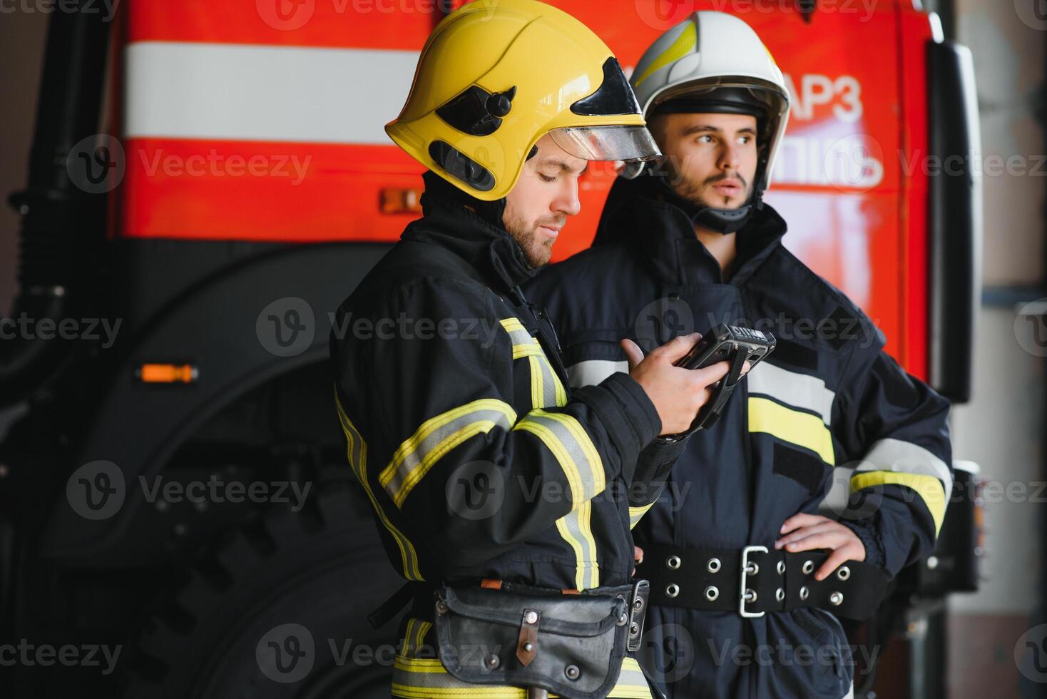 Two professional firefighters with uniforms and protective helmets posing infront of a firetruck. photo