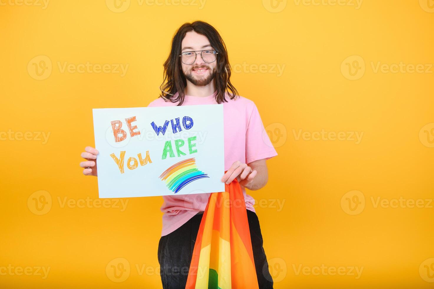 Young queer bisexual smiling gay man with make up in beige tank shirt hold card sign with be who you are title text on rainbow flag background studio portrait. photo
