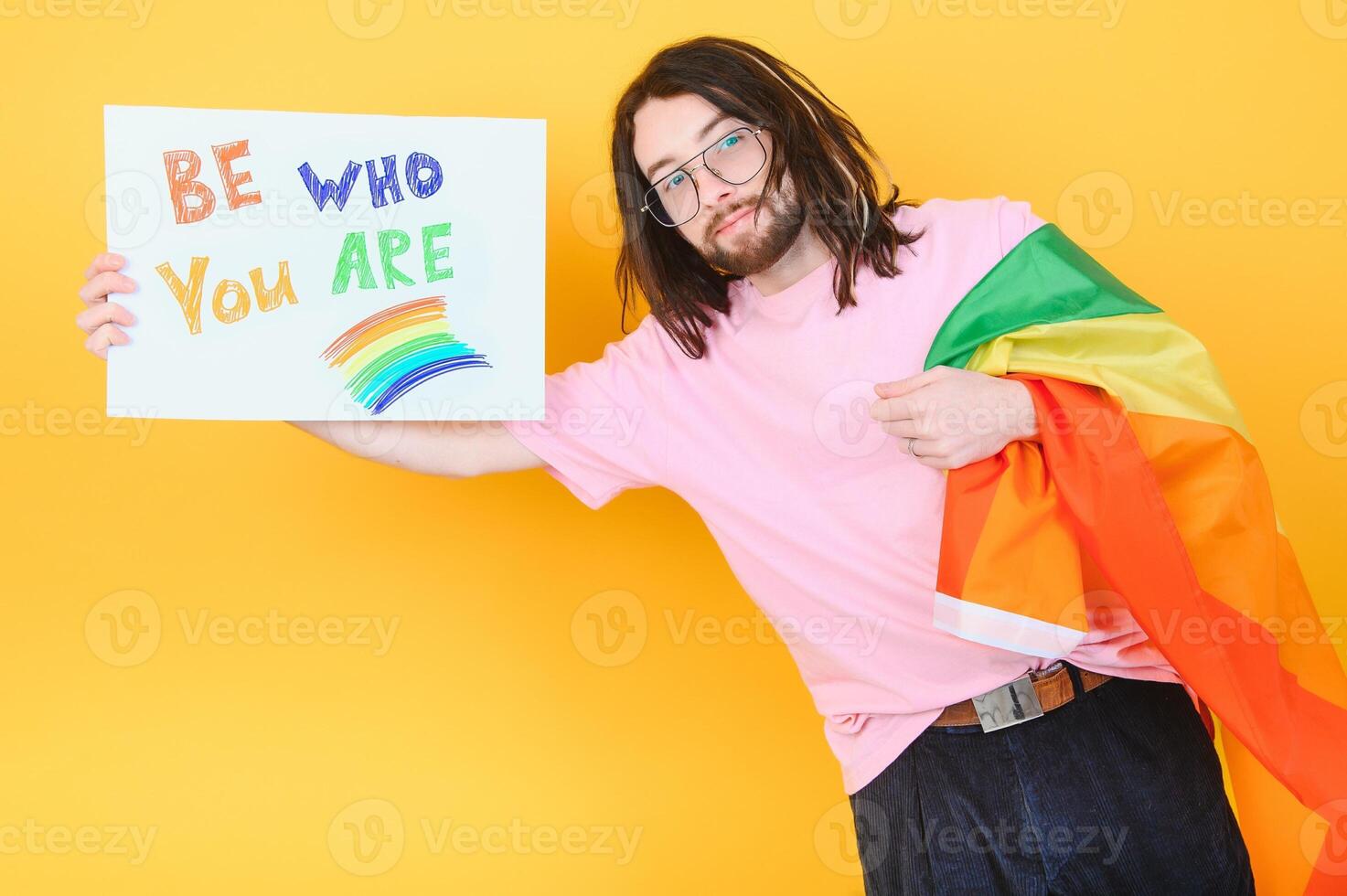 Young queer bisexual smiling gay man with make up in beige tank shirt hold card sign with be who you are title text on rainbow flag background studio portrait. photo