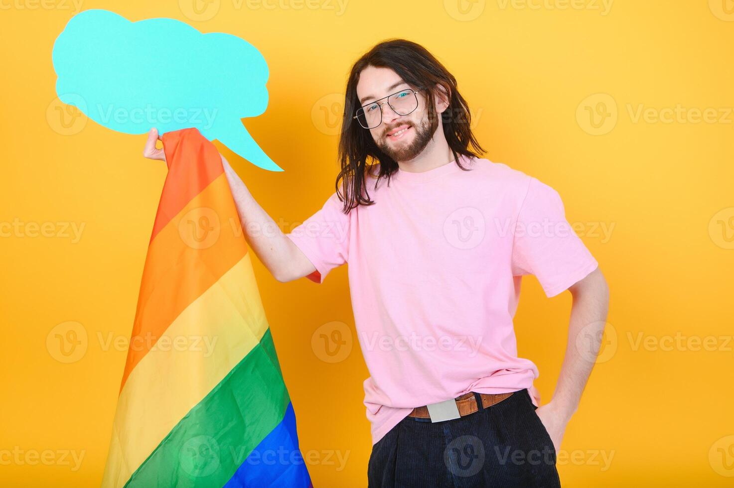 Portrait of cheerful smiling homosexual man holding empty blank board, with a gay pride flag at studio. Lgbtq flag, rainbow flag, celebrating parade. photo