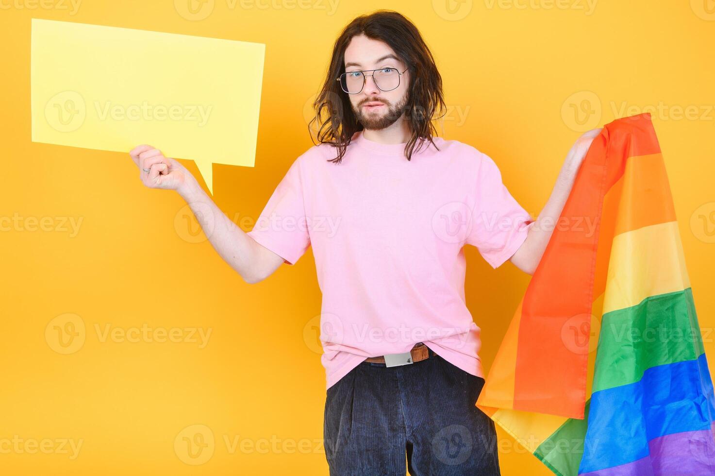 Portrait of cheerful smiling homosexual man holding empty blank board, with a gay pride flag at studio. Lgbtq flag, rainbow flag, celebrating parade. photo