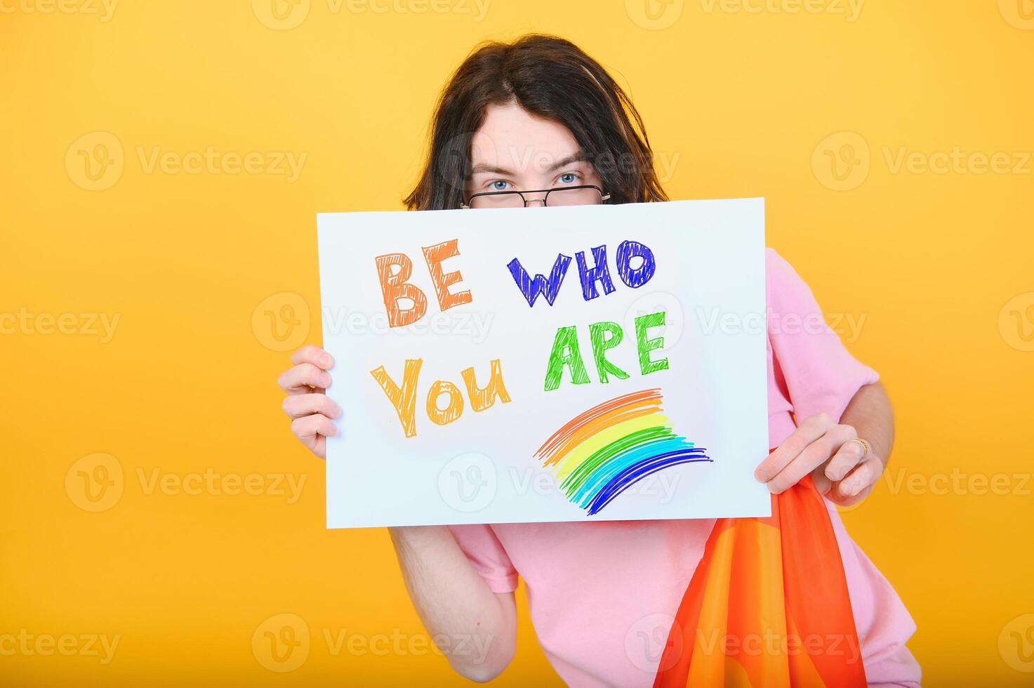 Young queer bisexual smiling gay man with make up in beige tank shirt hold card sign with be who you are title text on rainbow flag background studio portrait. photo