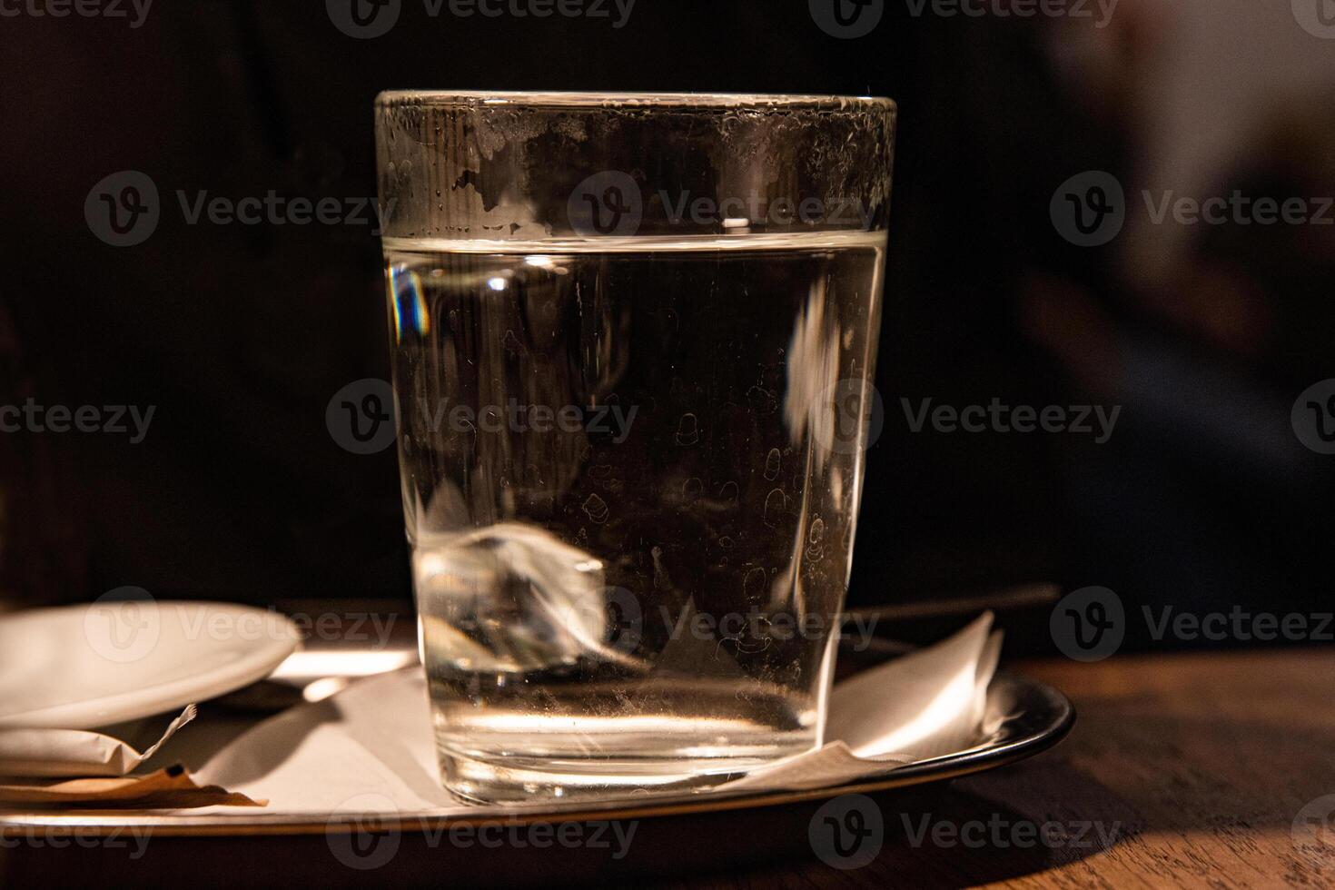 Close up of glass with hot water served on the white plate with napkin, sugar pack and tee bag on the wooden table in Cafe. Dark background. Drink concept. Tea cup. Selective focus. Copy space photo