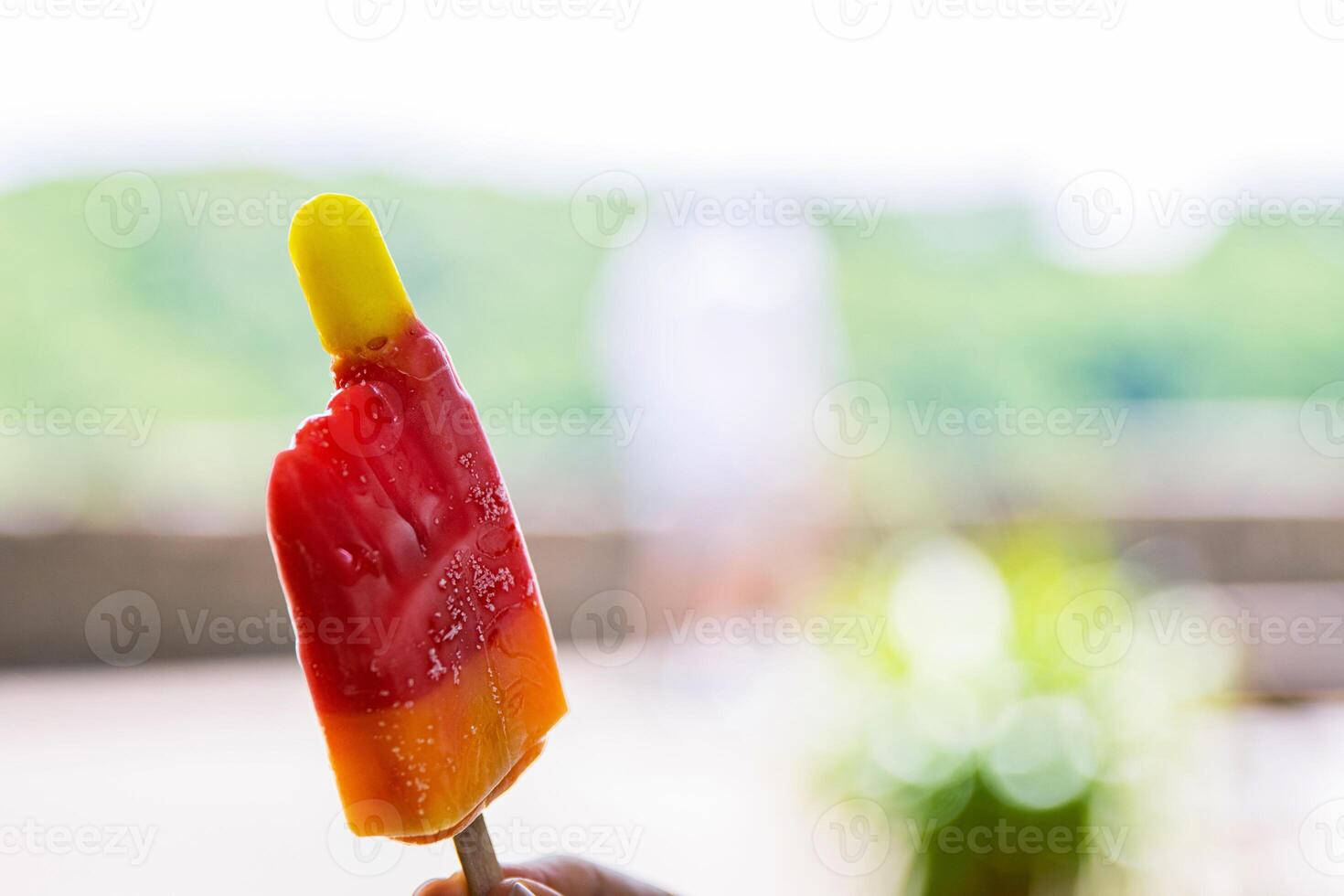 Girl holding and showing bitten yellow red ice cream her blurred face and silhouette are on background. Color frozen fruit popsicle. Hot Sunny day outside. Copy Space. Selective focus photo