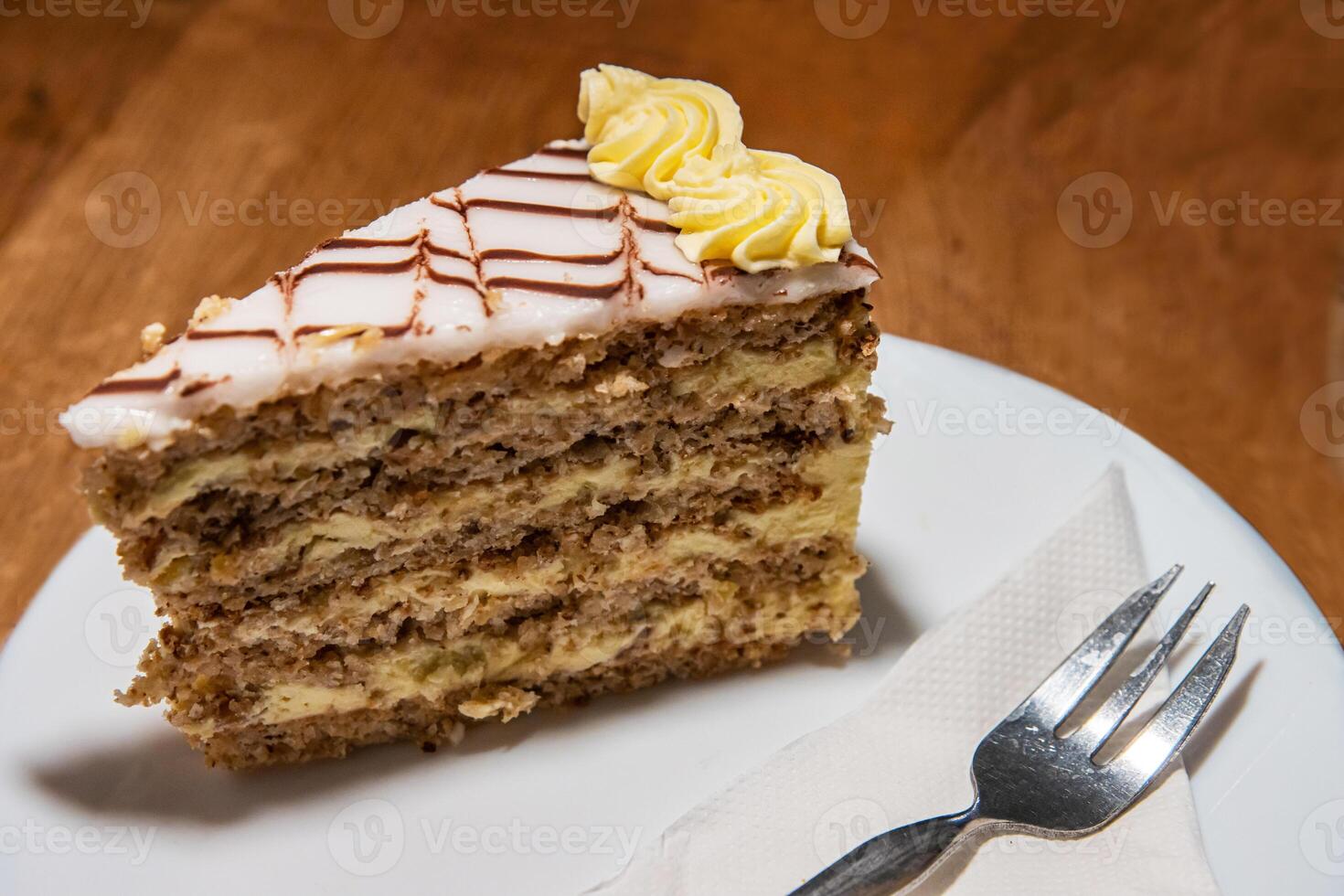 White plate with slice of tasty homemade delicious sweet Esterhazy cake with nuts and cream. Piece of tasty Esterhazy Torte served with napkin and fork. Wooden table in cafe shop. Selective focus photo