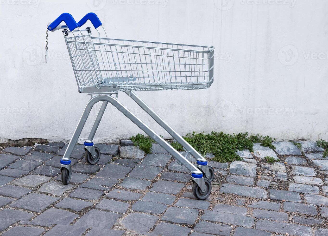 Abandoned empty shopping metal cart with blue grip standing in the store backyard by dirty concrete wall on the cobblestone road. Old dirty grunge walls. Lost place. Copy space. Selective focus. photo