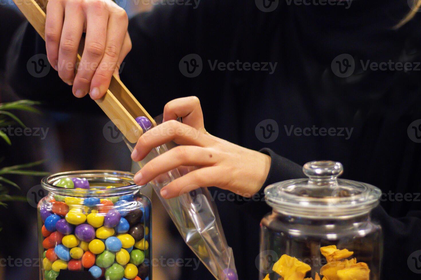 Young woman selling candies at the food market. She is picking out candy with wooden or bamboo tongs out of the glass jar and putting them into plastic bag. Copy space. Selective focus, Blurred hands photo