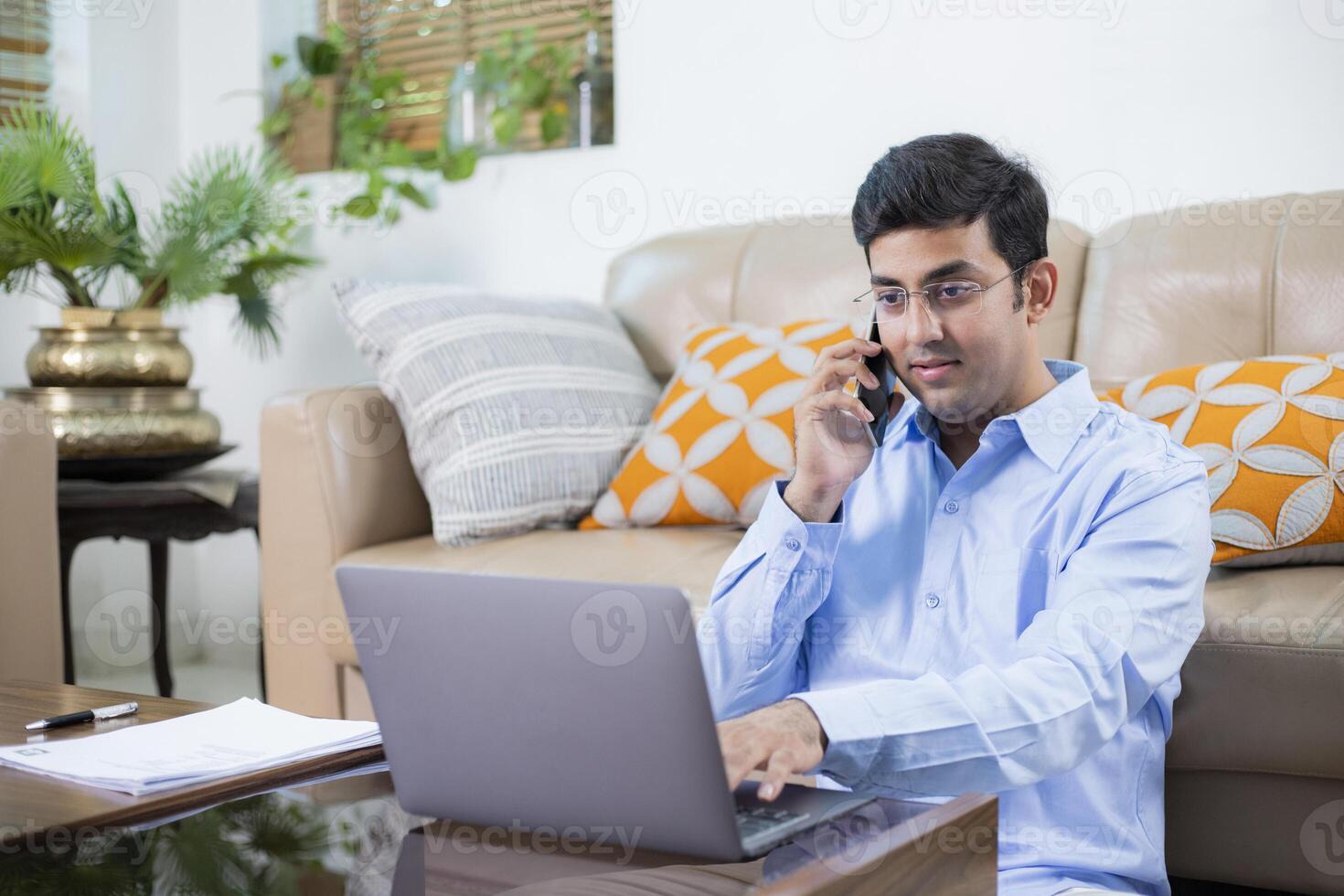 Man In Blue Shirt Working On Laptop Pointing At Screen While Discussing On Mobile Phone photo